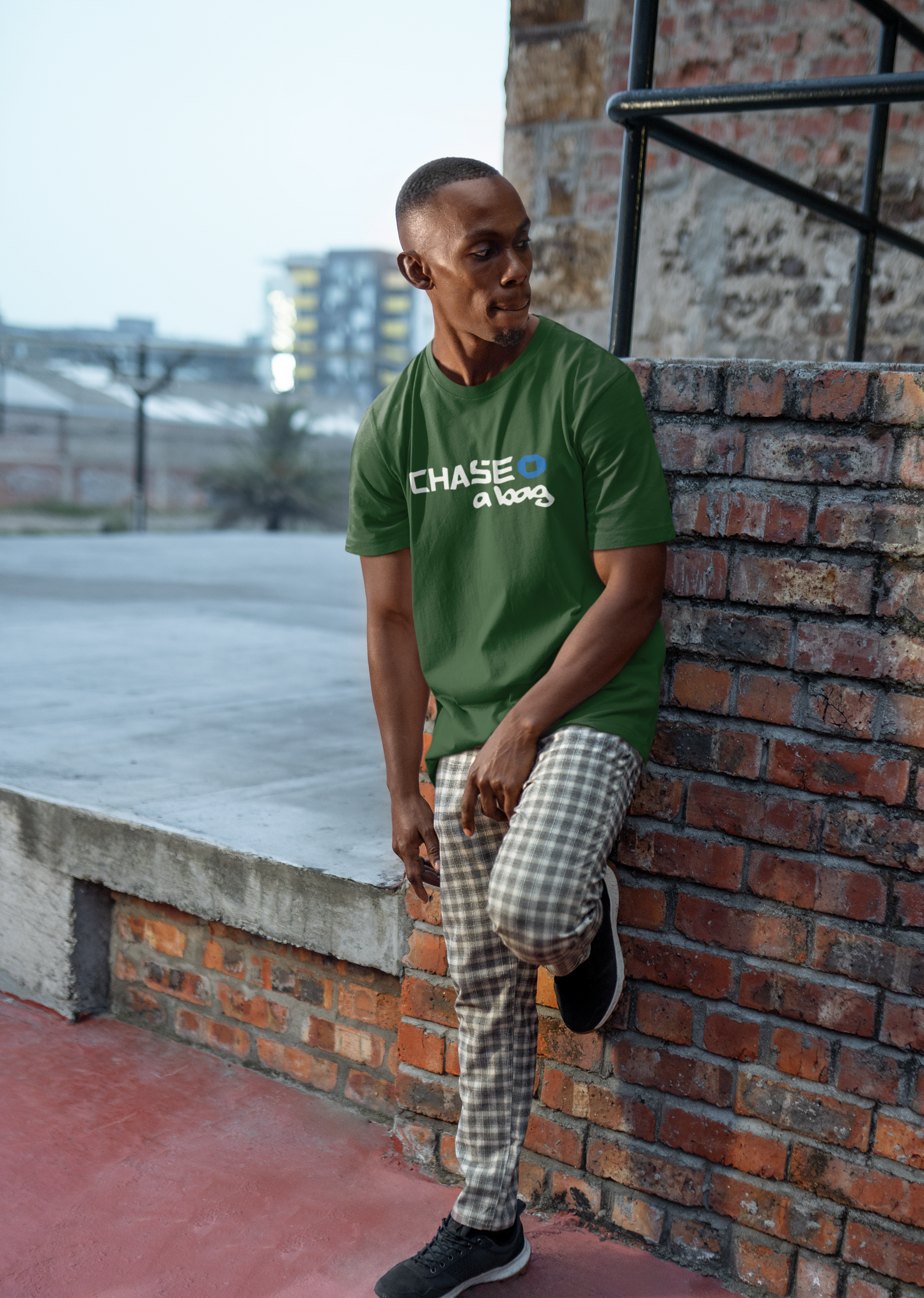 A person wearing a yellow t-shirt with the text 'CHASE A BAG' printed on the front, standing in front of a brick wall and a building.